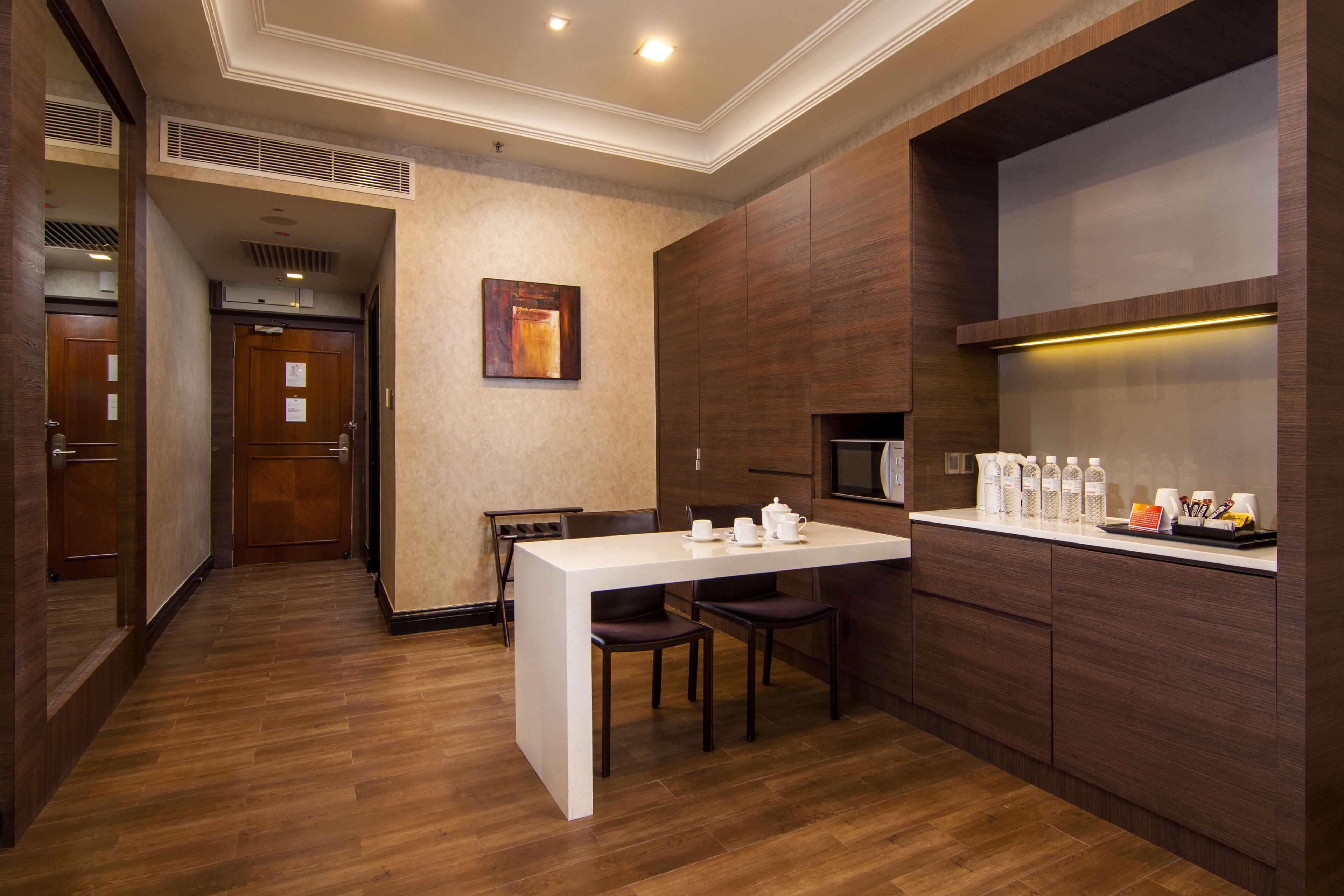 Una habitación de hotel moderna con suelo de madera, un escritorio blanco con dos sillas, un microondas y una pequeña kitchenette.