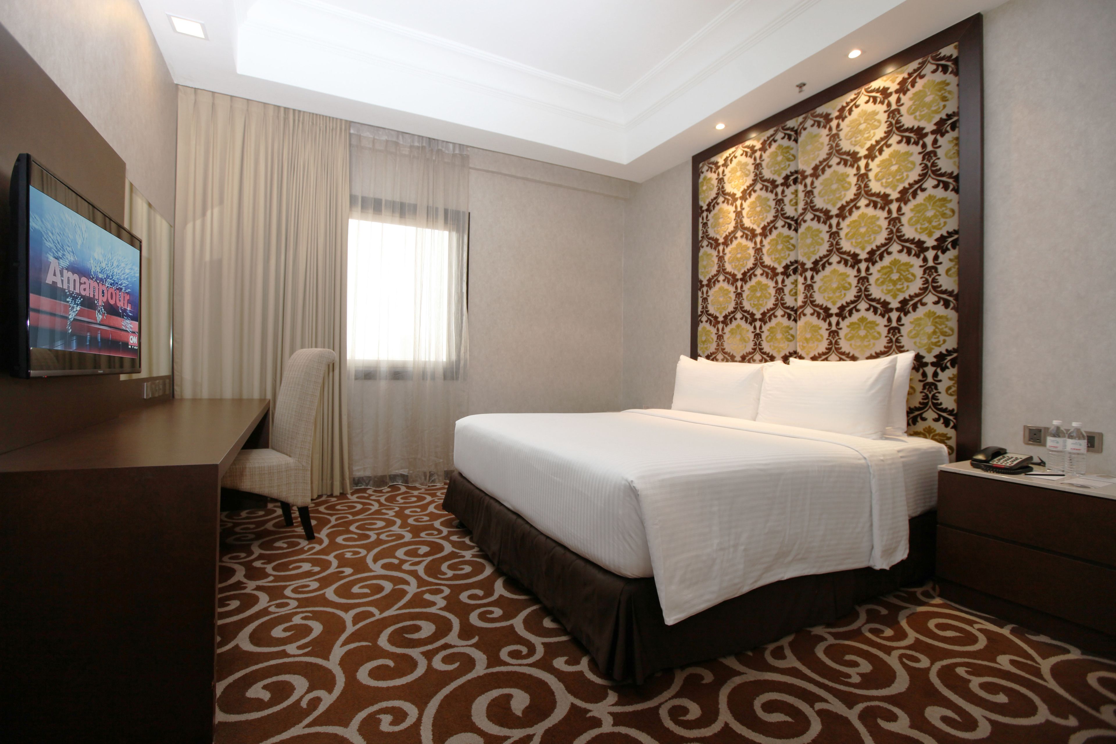 Habitación de hotel con una cama grande, un escritorio, una silla, un televisor y una alfombra estampada.