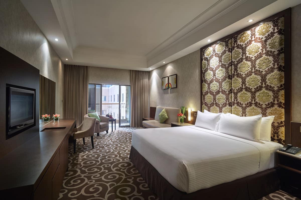 Ein Hotelzimmer mit einem großen Bett, einem Schreibtisch mit Fernseher, einem Sessel, einer Schreibtischlampe und einem gemusterten Teppich.