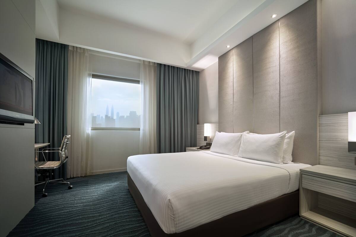 Ein Hotelzimmer mit einem großen Bett, einem Schreibtisch, einem Sessel, einem Fernseher und Blick auf die Stadt.