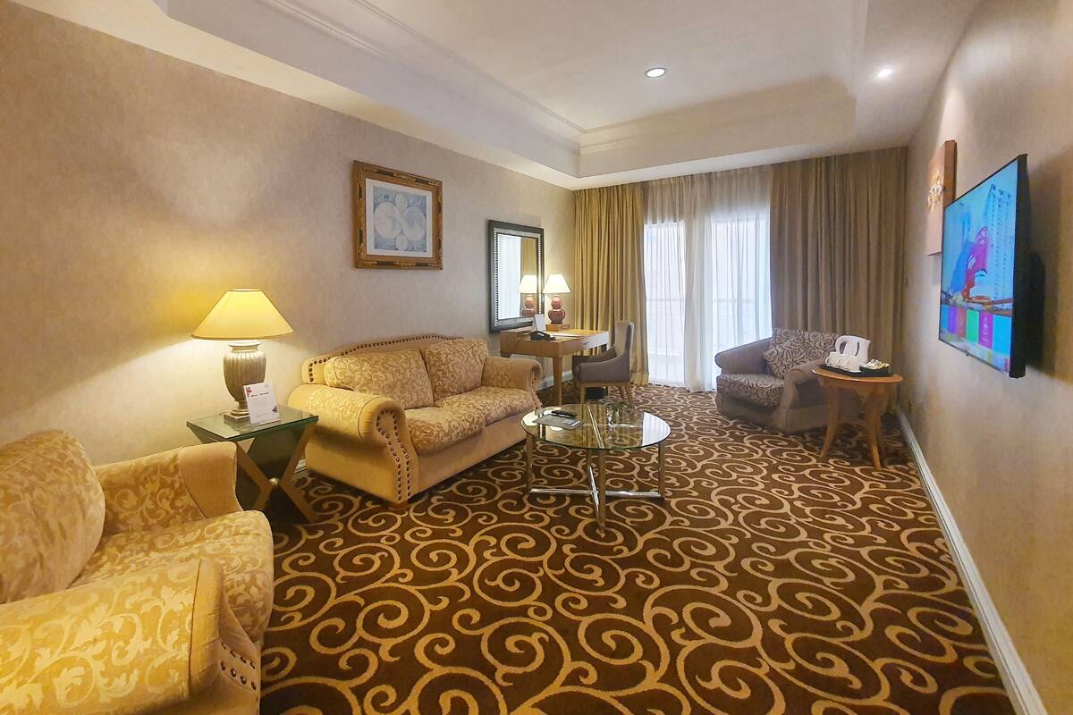 Ein Hotelzimmer mit Teppichmuster, einer Couch, einem Sessel, einem kleinen Tisch, einer Lampe, einem Fernseher und einem fenster mit Vorhängen.