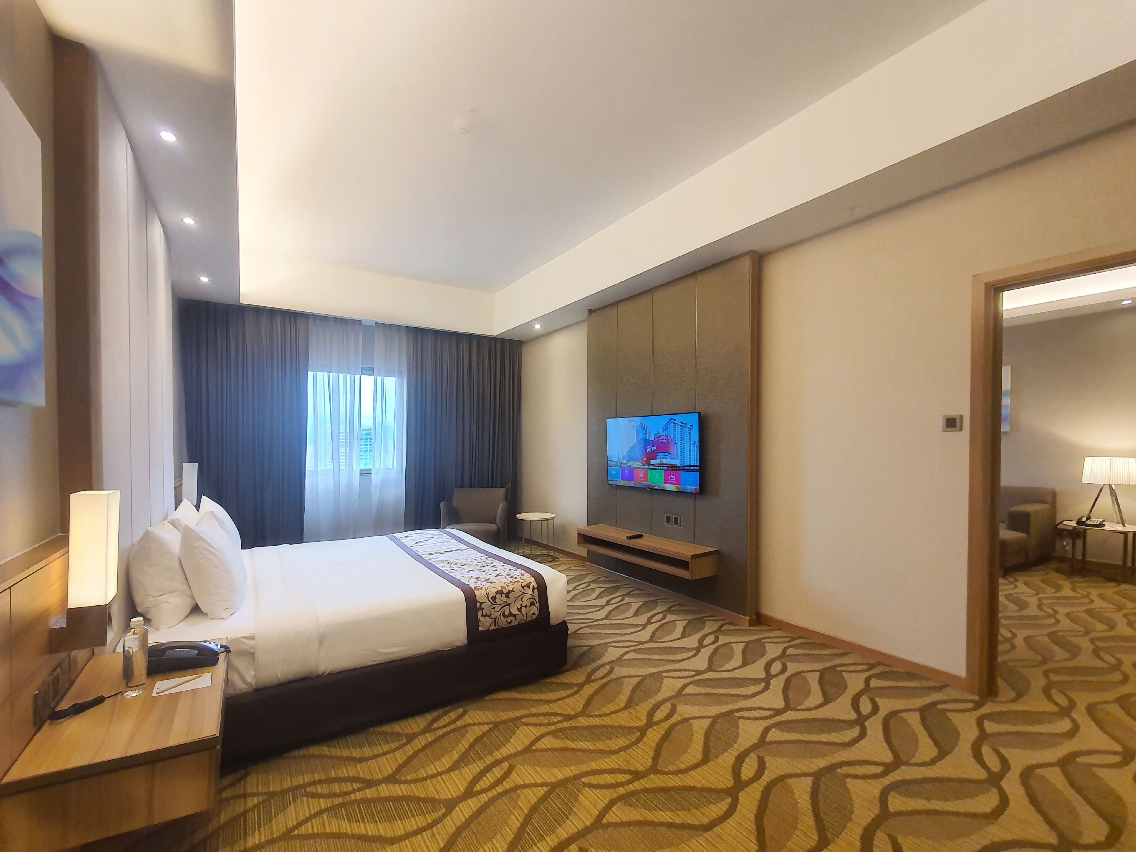 Habitación de hotel con una cama grande, un televisor montado en la pared, una mesita con una lámpara y una alfombra estampada.