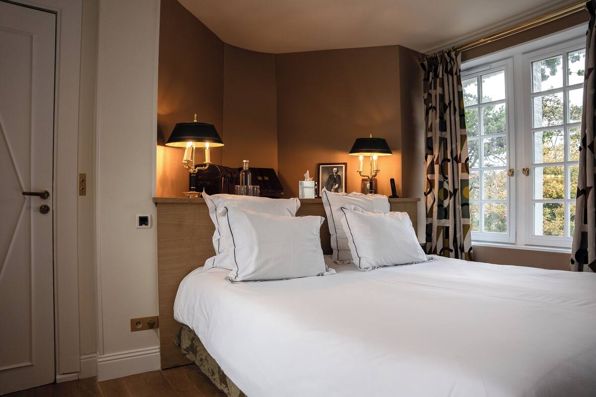 A neatly made bed with white linens and pillows, two bedside lamps, a framed picture, and a window with curtains.