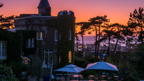 Un bâtiment historique aux murs couverts de lierre, doté d’une tourelle et offrant une vue sur le coucher de soleil sur la mer.