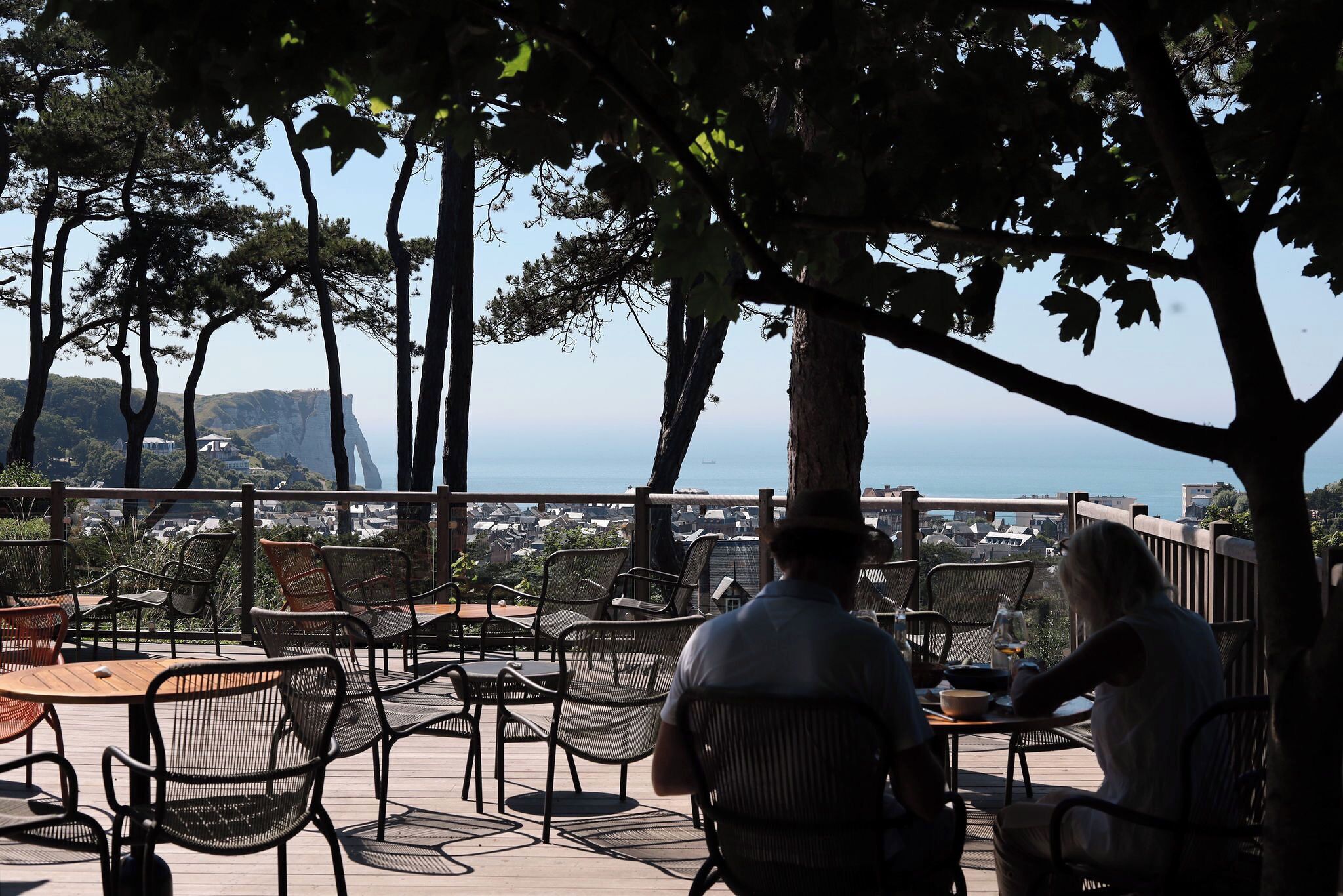 An outdoor seating area with tables and chairs, overlooking a coastal landscape with trees and a view of the sea.