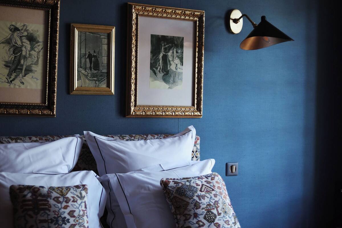 A neatly made bed with patterned pillows, a wall-mounted lamp, and framed artwork on a dark blue wall.