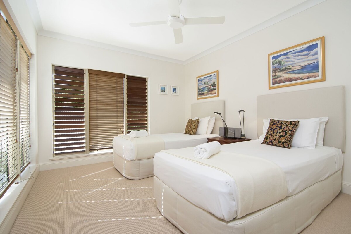 A hotel room with two beds, a ceiling fan, large windows with blinds, and artwork on the walls.