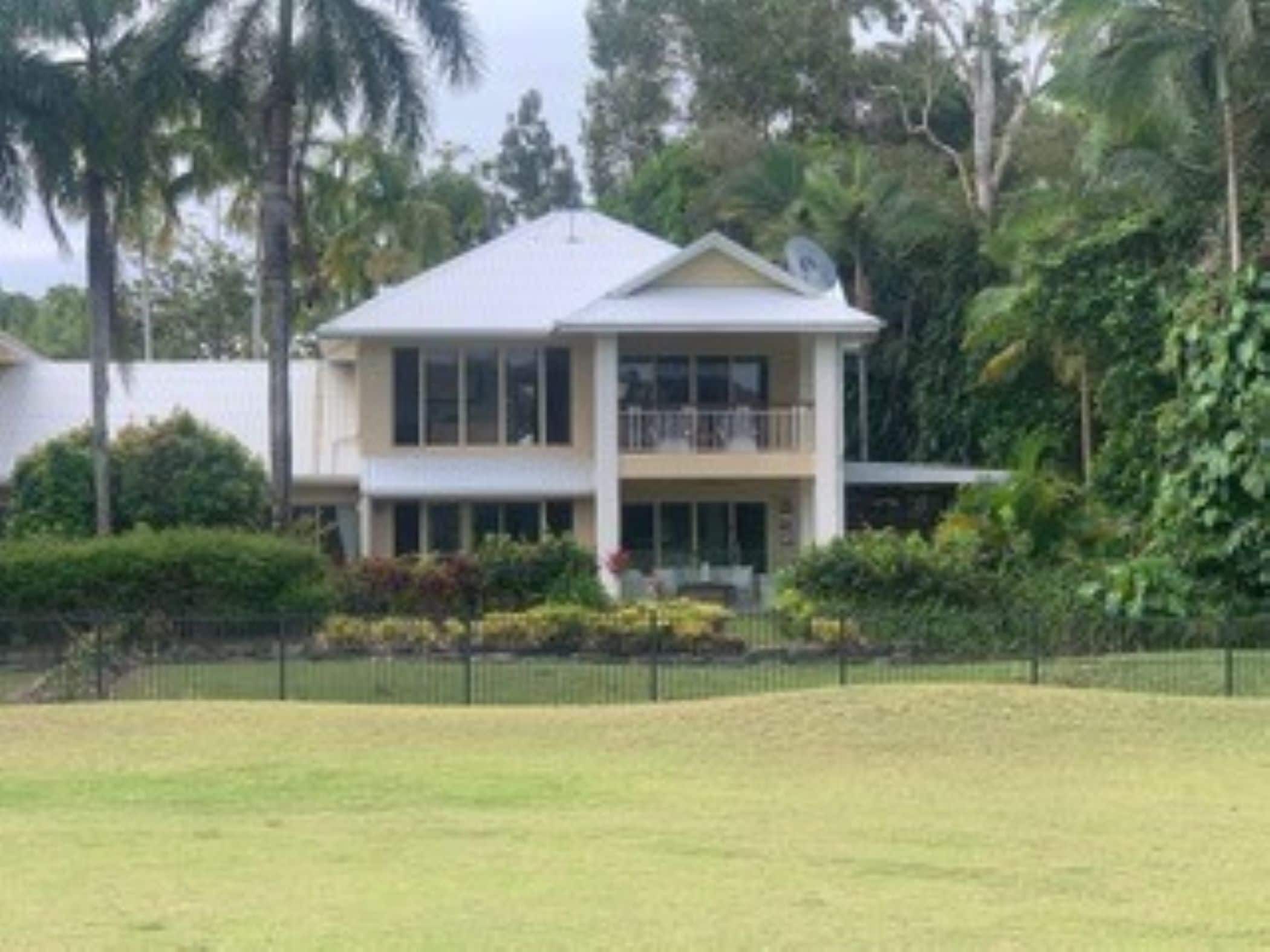 A two-story house with a balcony, surrounded by lush greenery and a fenced yard.