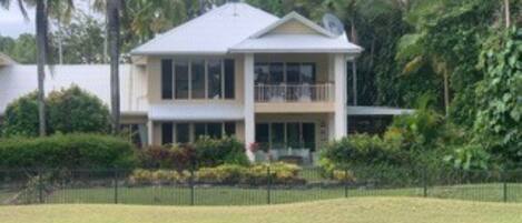 A two-story house with a balcony, surrounded by lush greenery and a fenced yard.