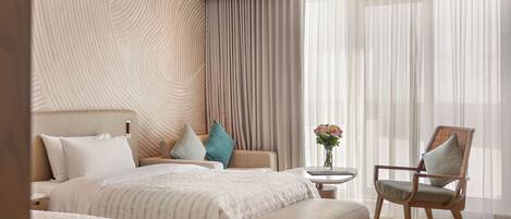 Une chambre d’hôtel moderne avec un grand lit, un coin salon comprenant un fauteuil et un pouf, une petite table ornée d’un vase de fleurs et une grande fenêtre avec des rideaux légers.