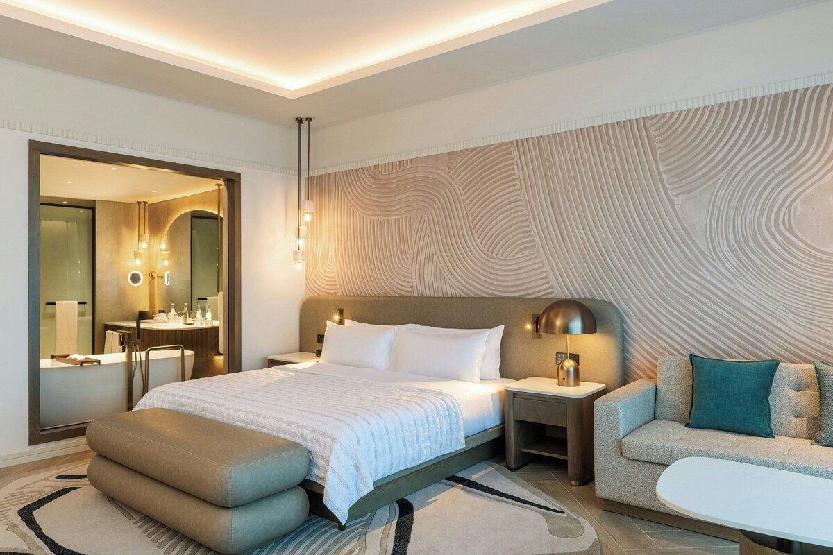 A modern hotel room with a large bed, bedside tables, a sofa, and a bathroom with a mirror and lights.