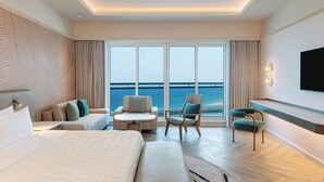 A modern hotel room with a large bed, a sofa, a chair, a small table, a TV, and a view of the sea through large windows.