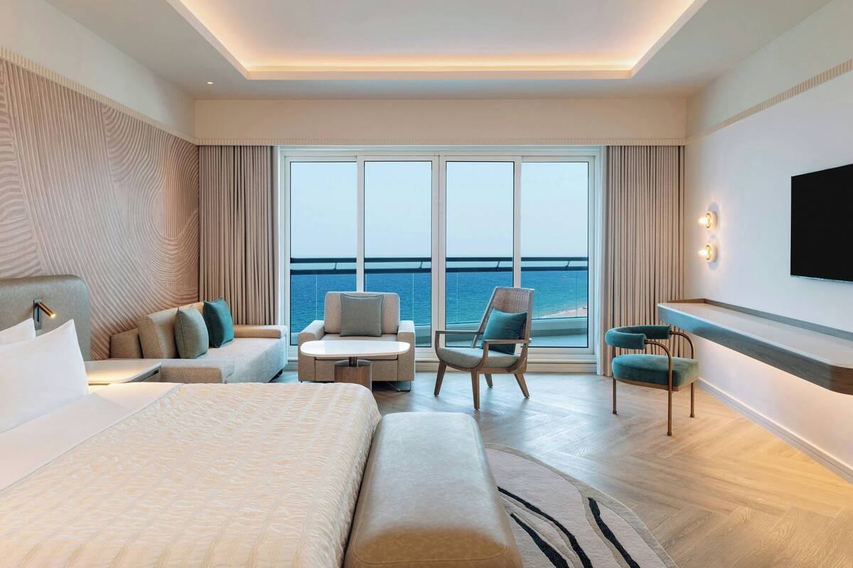 A modern hotel room with a large bed, a sofa, a chair, a small table, a TV, and a view of the sea through large windows.