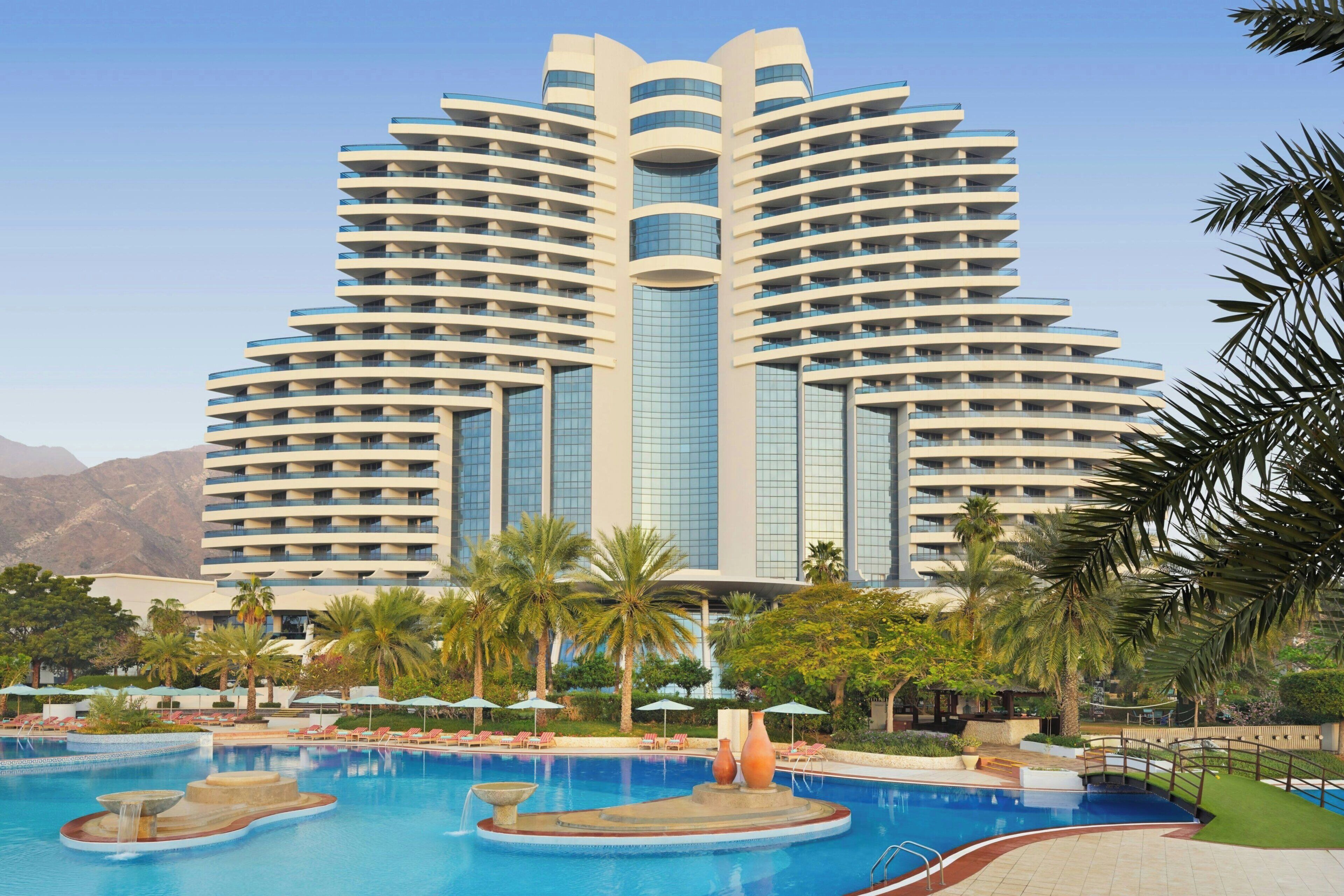 Een modern hotel met een centraal gelegen zwembad, meerdere balkons en palmbomen.