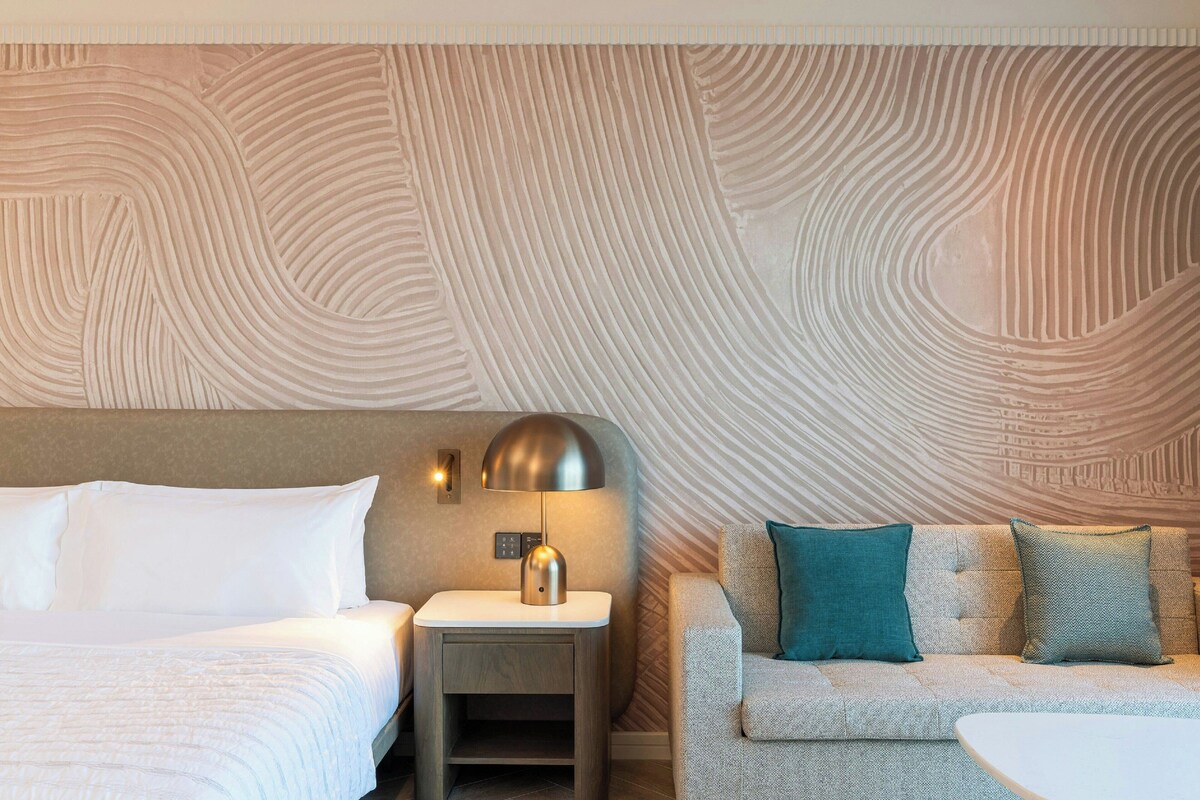 Quarto de hotel com uma cama, uma mesa de cabeceira com abajur, um sofá e um papel de parede estampado.