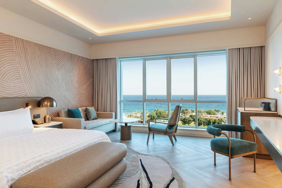 A modern hotel room with a large bed, a sofa, two chairs, a desk, and a view of the ocean.