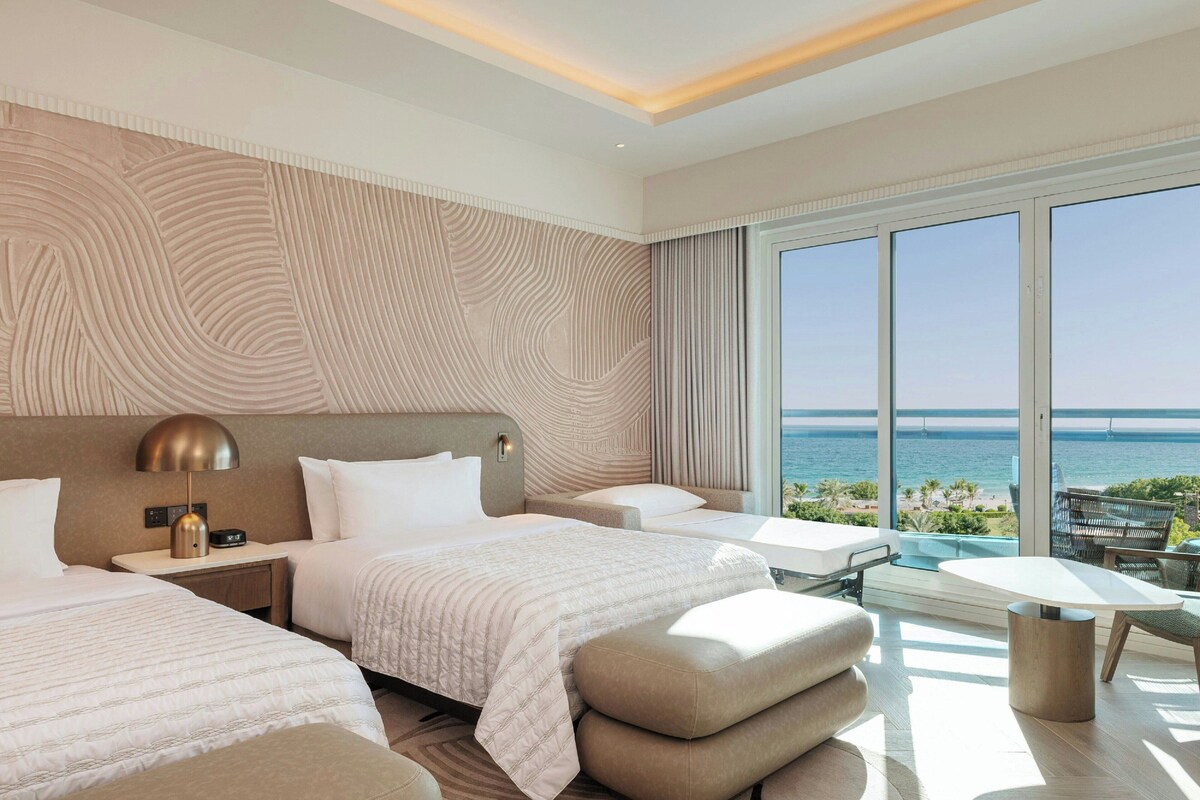 Quarto de hotel com duas camas, uma janela ampla com vista para o mar, varanda com mesa e cadeiras, e design moderno com parede texturizada.