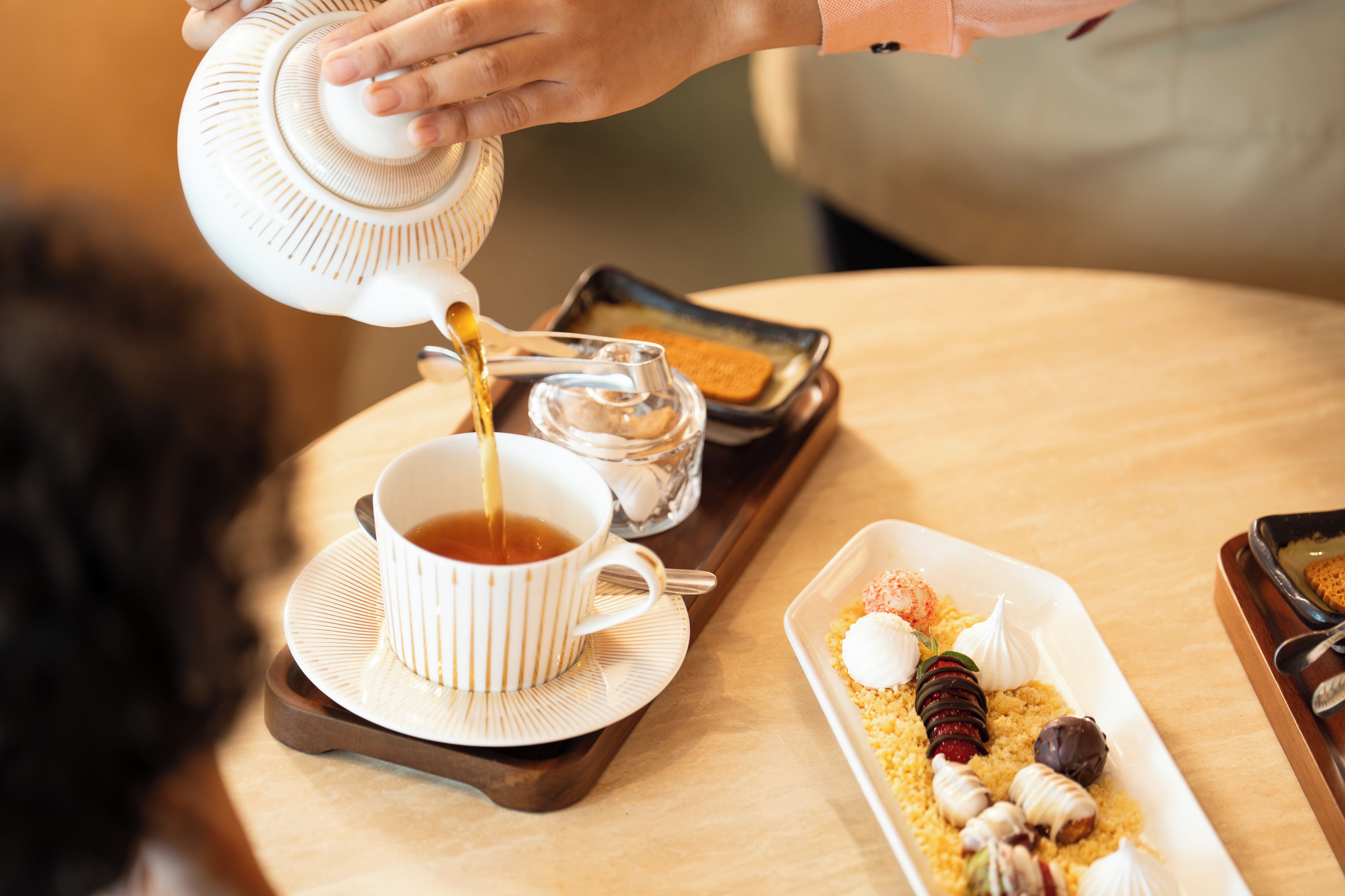 Une personne verse du thé dans une tasse sur un plateau, à côté d’une assiette de dessert.