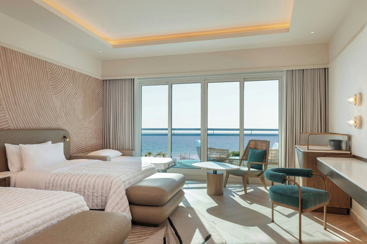 Quarto de hotel com uma cama grande, uma escrivaninha, uma cadeira e vista para o mar.