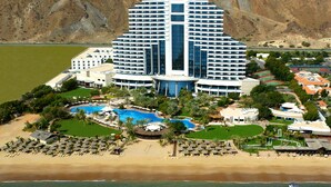 Un gran hotel con una piscina central, varios balcones y una zona de playa con tumbonas.