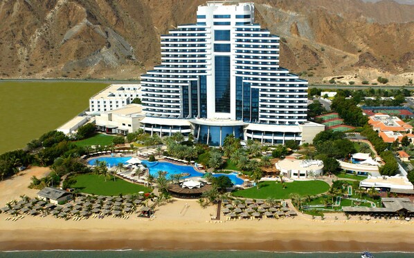 Um hotel grande com piscina central, várias varandas e área de praia com espreguiçadeiras.