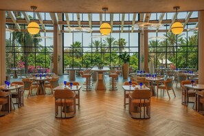 Un amplio comedor interior con varias mesas y sillas, ventanas grandes que ofrecen vistas a palmeras y flores, y lámparas esféricas colgantes.