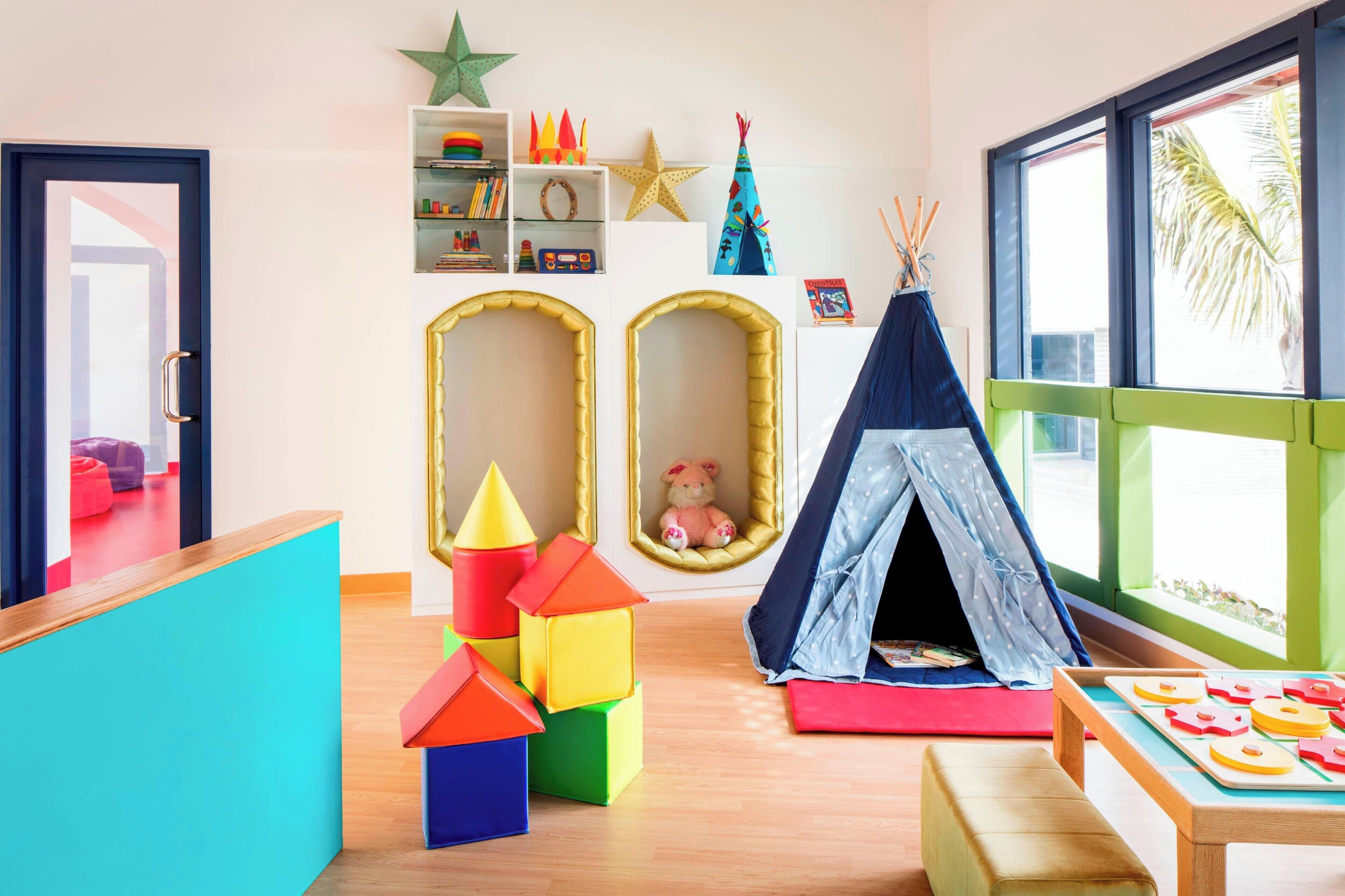 Une salle de jeux pour enfants avec des blocs de construction colorés, une tente et une fenêtre donnant sur des palmiers.