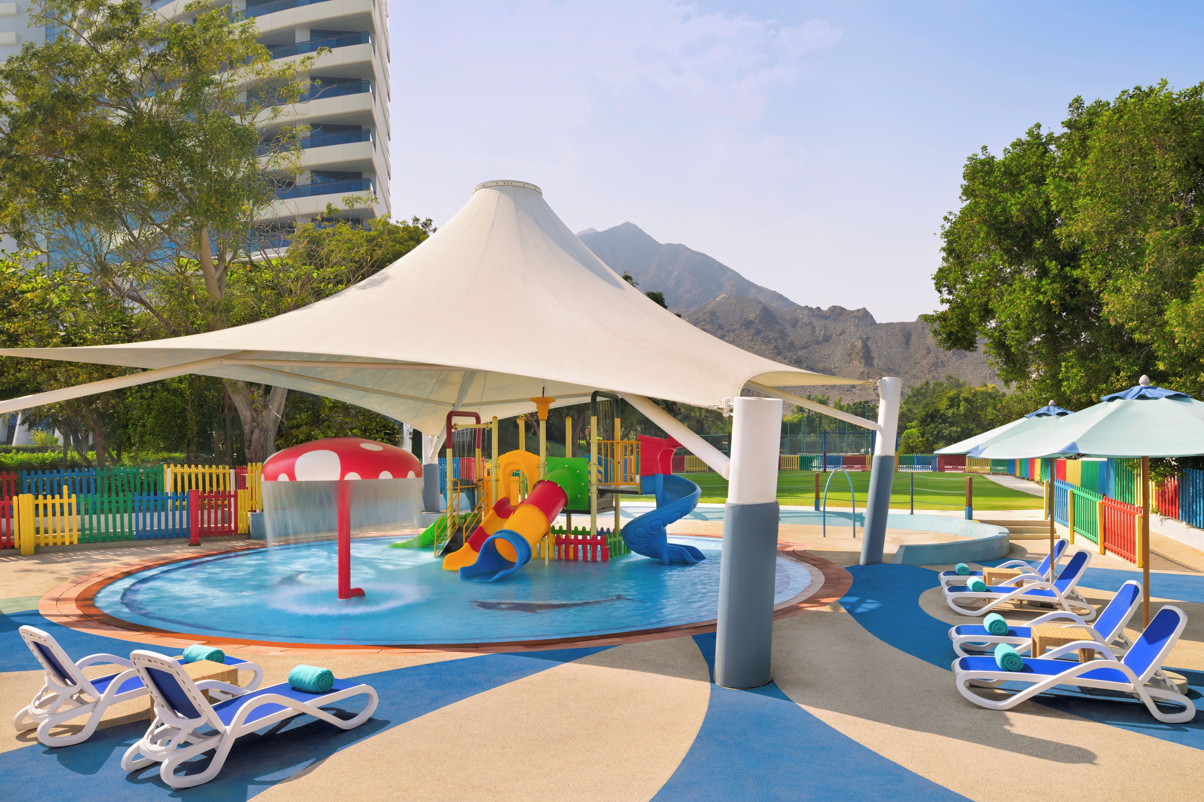 Un área de juegos infantiles con toboganes y piscina, rodeada de sillones de descanso y una sombrilla grande.