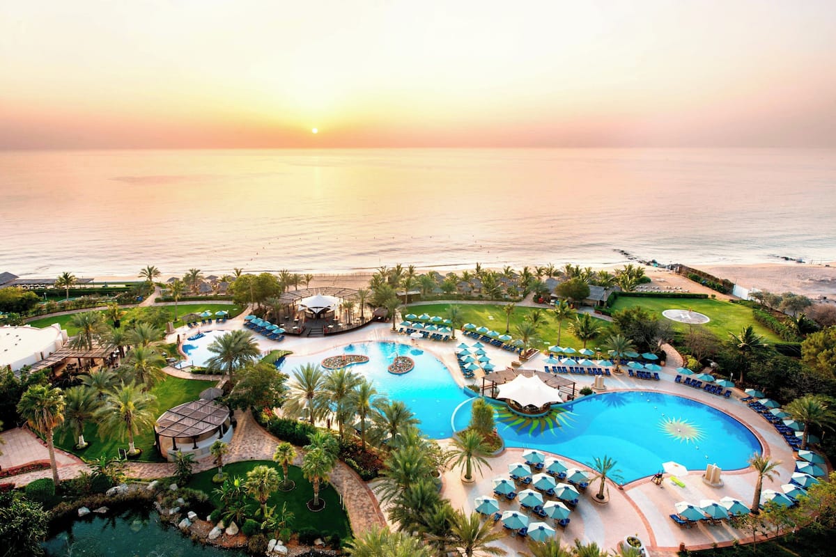 A resort with multiple swimming pools, palm trees, and a view of the ocean at sunset.