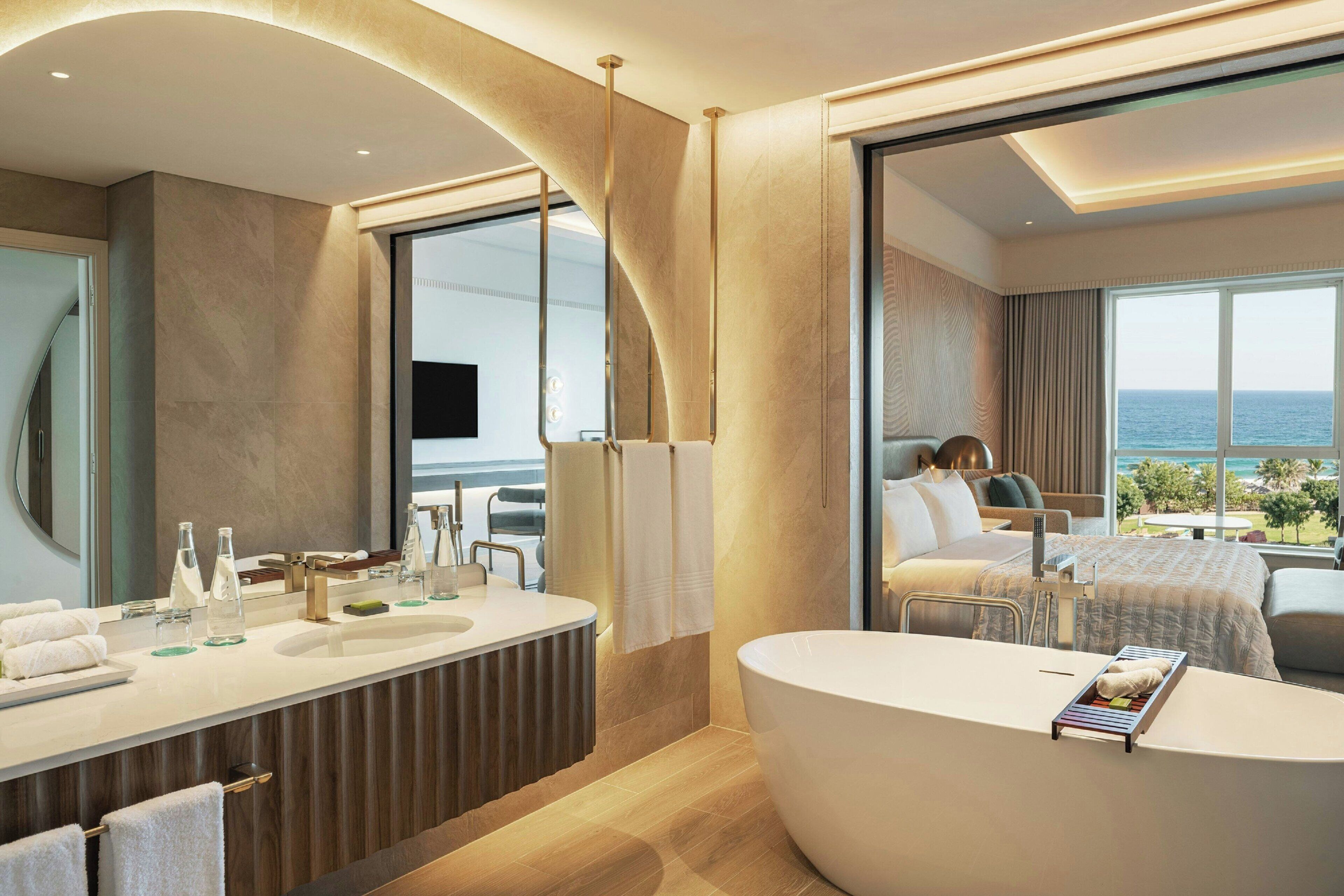 A modern bathroom with a large bathtub, a vanity with a mirror, and a large window offering a view of the sea.