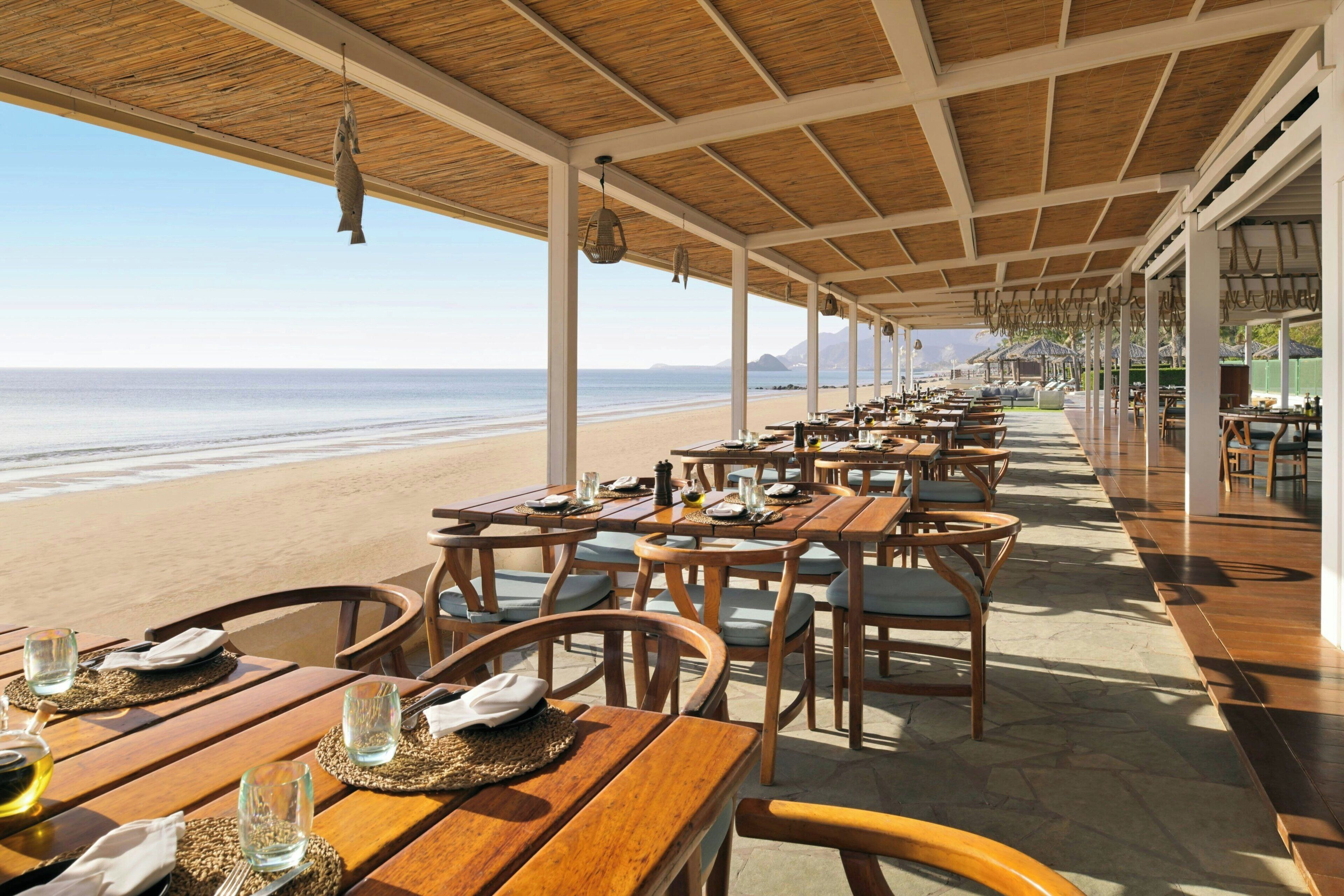 Un área de comedor al aire libre con mesas y sillas, con vista a una playa.
