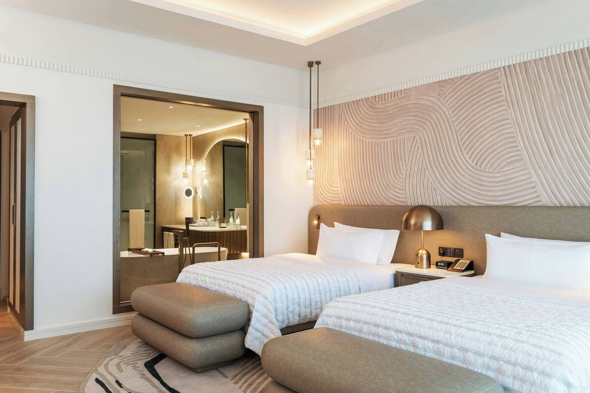 A hotel room with two beds, a large mirror, a vanity with a sink, and a modern light fixture.