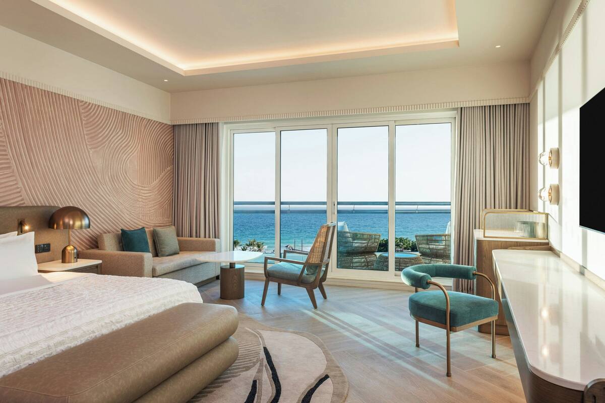 A modern hotel room with a large bed, a sitting area with a view of the sea, a dining table, and a TV.