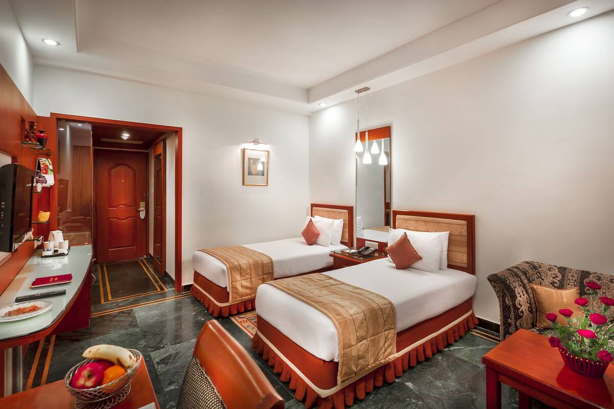 Habitación de hotel con dos camas, un escritorio, una silla, una mesita pequeña, un espejo y un jarrón decorativo con flores.