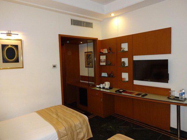 A hotel room with a bed, a desk with a flat-screen TV, a coffee maker, and a shelf with decorative items. - Regency Madurai by GRT Hotels (Madurai)
