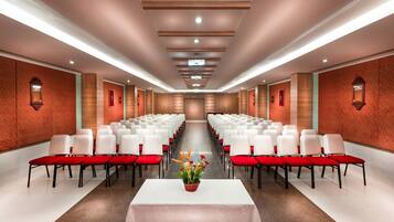 A conference room with rows of white chairs and red cushions, a central table with a floral arrangement, and a red carpeted floor.