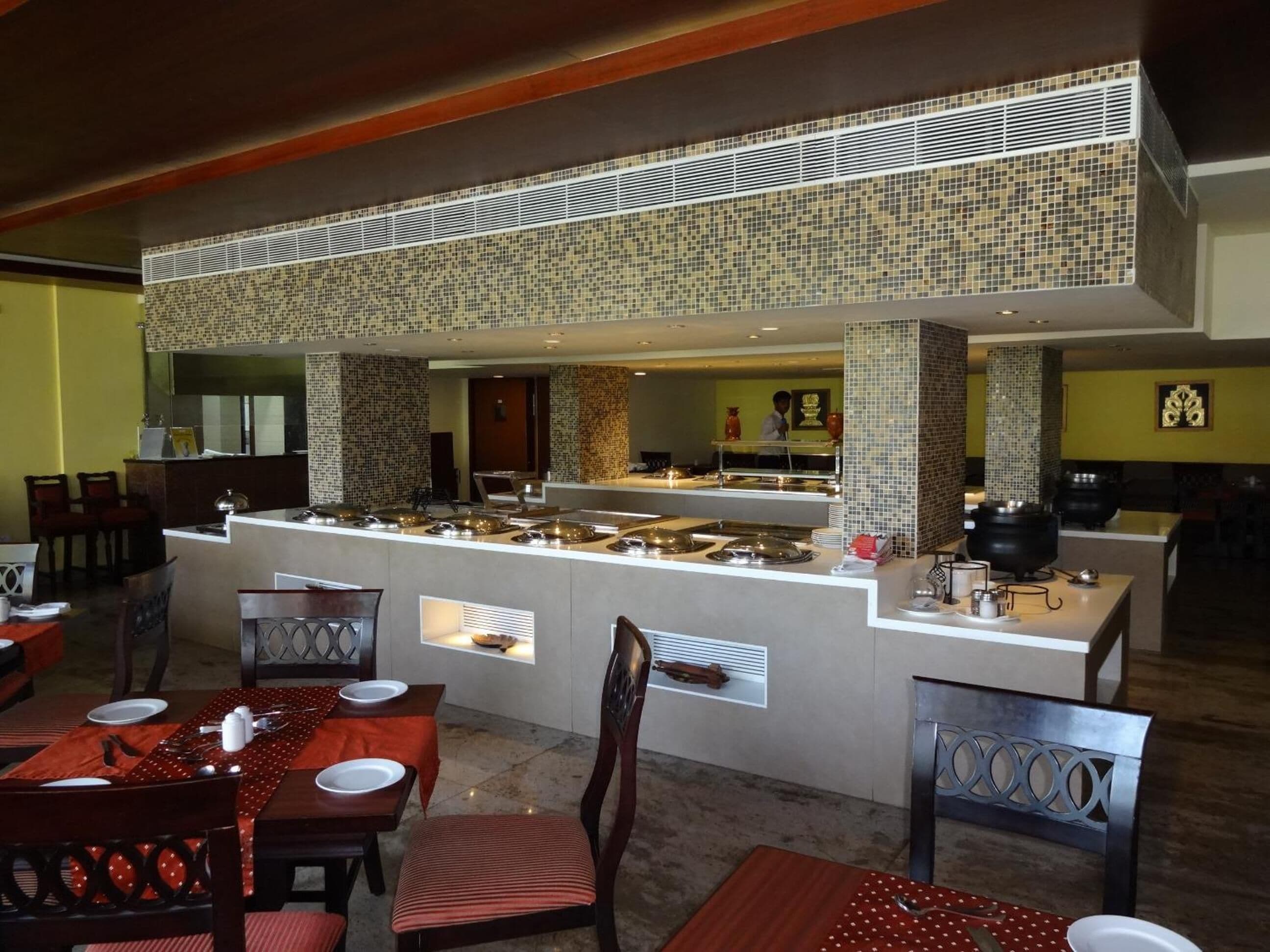 Un área de bufé con varias estaciones de servicio, una zona de estar con mesas y sillas, y una cocina visible al fondo.