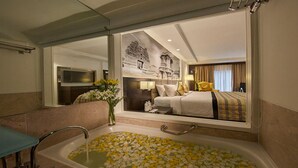 Ein Hotelzimmer mit einem großen Bett, einem Badezimmer mit einer Badewanne voller Blütenblätter, einem Spiegel und einem an der Wand befestigten Fernseher.