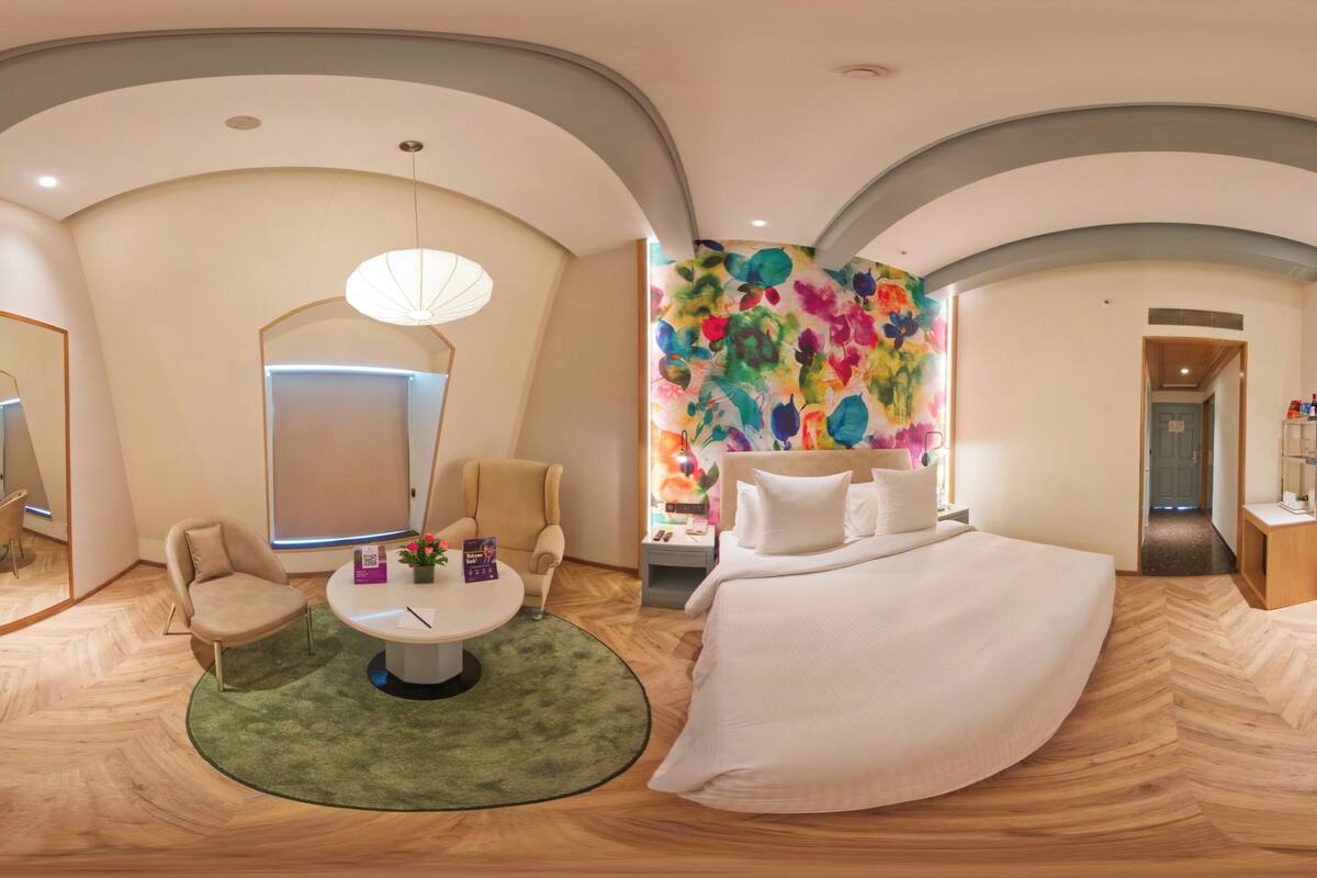 Een moderne hotelkamer met een groot bed, een ronde tafel met een groen vloerkleed, kleurrijke abstracte wandkunst en een grote spiegel.