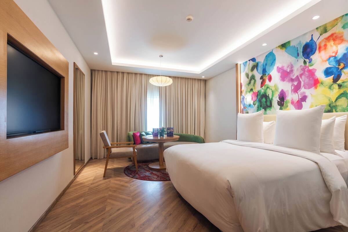 Een moderne hotelkamer met een groot bed, een flatscreen televisie aan de muur, een groene bank, een klein tafeltje met een vaas, en kleurrijke bloemenkunst aan de wand.
