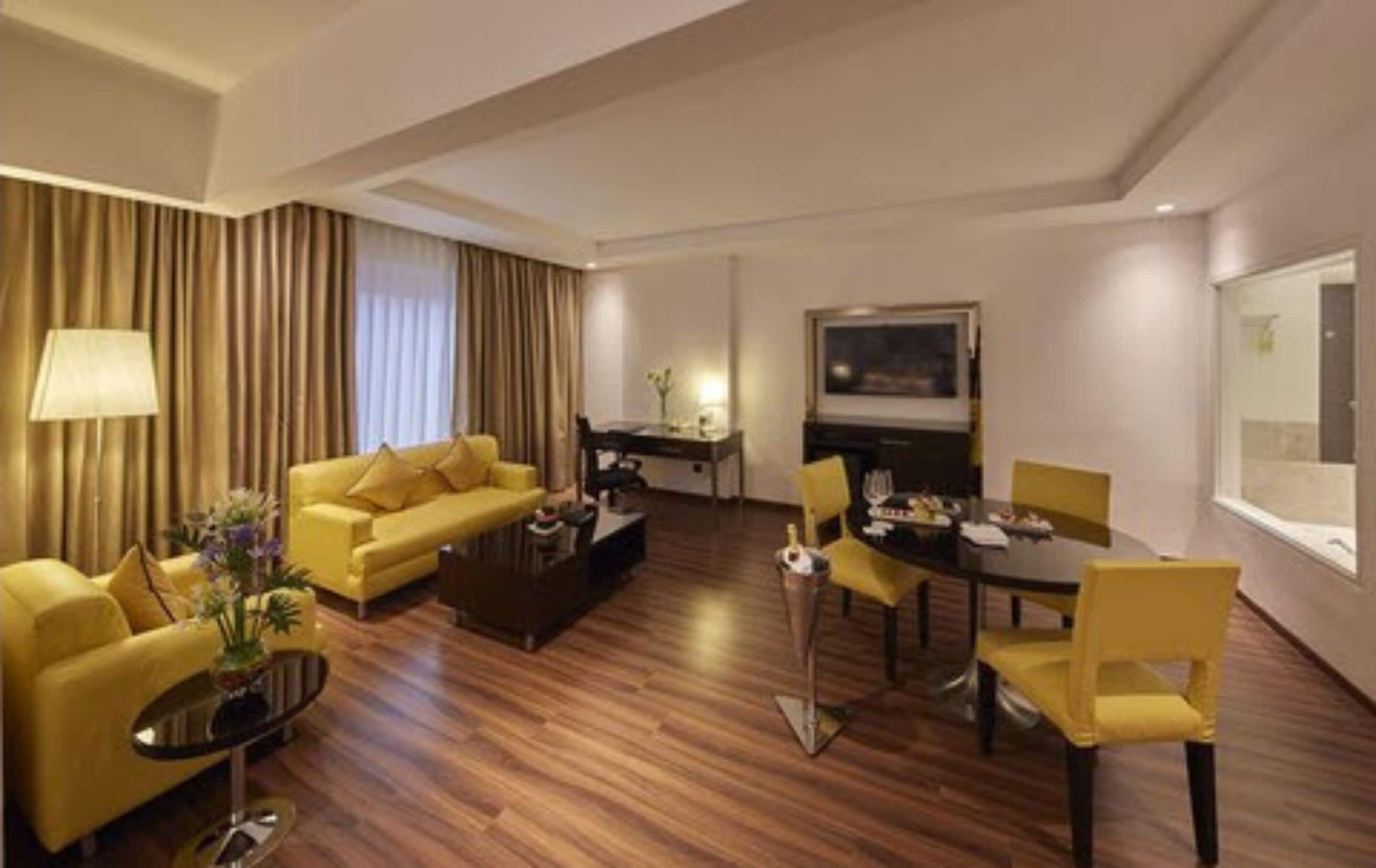 A hotel room with a yellow sofa, a dining table with chairs, a coffee table, a TV, and a lamp.