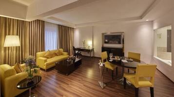 A hotel room with a yellow sofa, a dining table with chairs, a coffee table, a TV, and a lamp.
