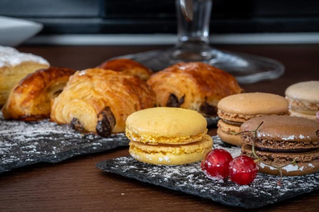 A variety of pastries on a slate serving board, including croissants and macarons, with a glass of wine in the background.