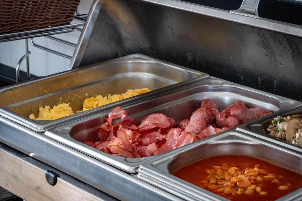 Un bufé con diferentes opciones de comida, incluyendo huevos revueltos, tocino, salchichas y frijoles en salsa.