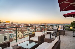 A rooftop terrace with wicker furniture, a glass table, and a view of the cityscape.