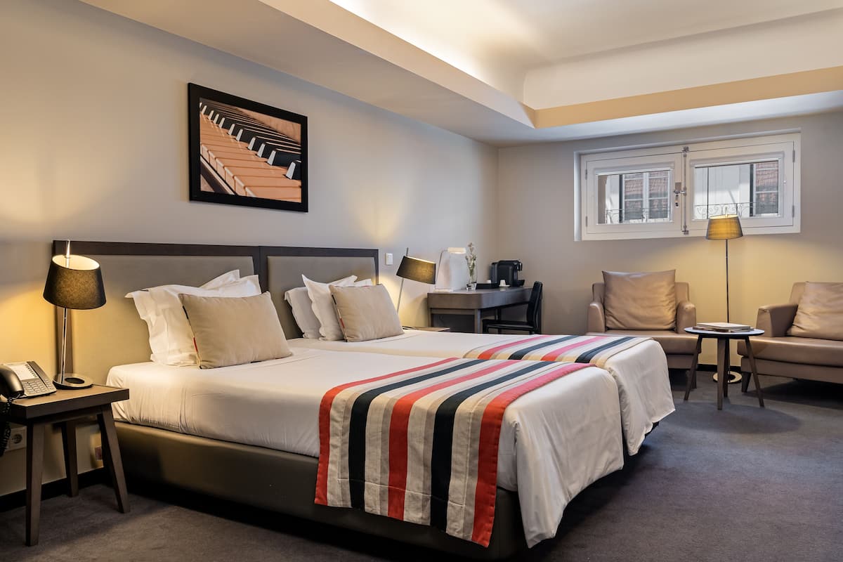 Habitación de hotel con una cama grande, dos mesitas de noche, un escritorio, una silla y un ventanal con vistas a edificios.