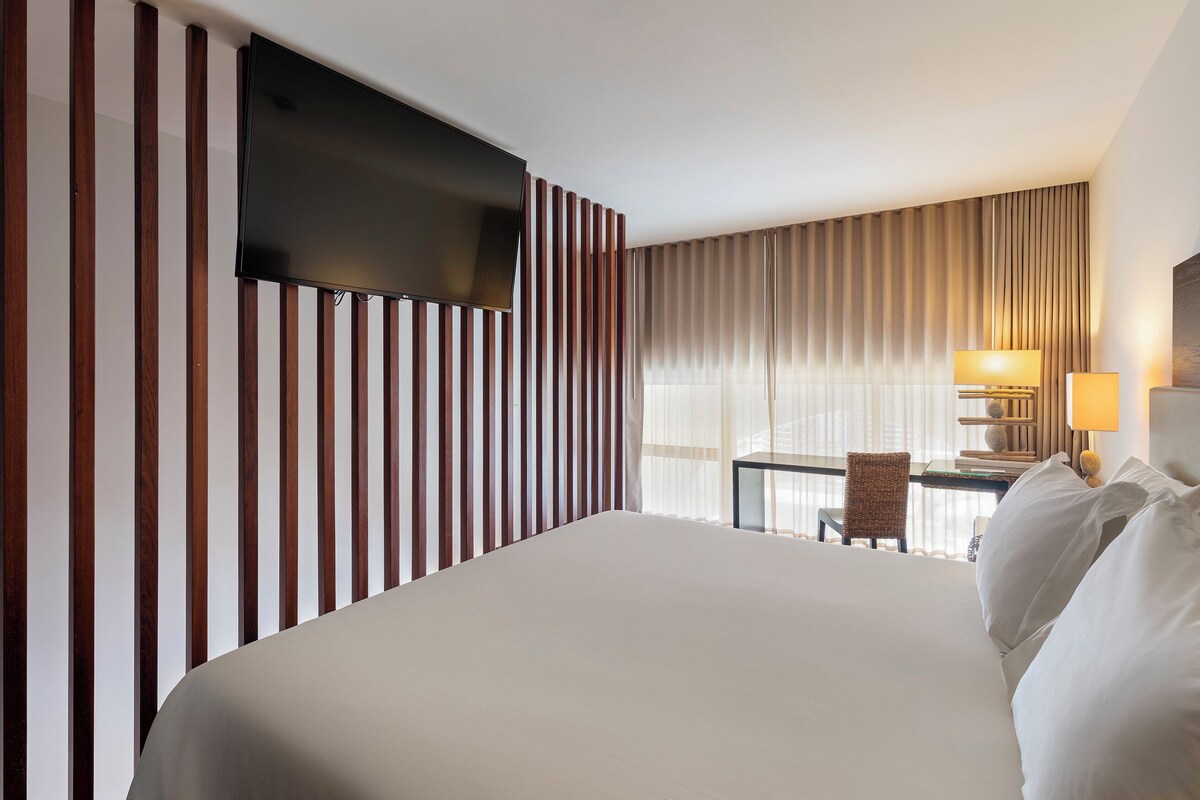 A hotel room with a large bed, a flat-screen TV mounted on the wall, a desk with a chair, and a lamp.