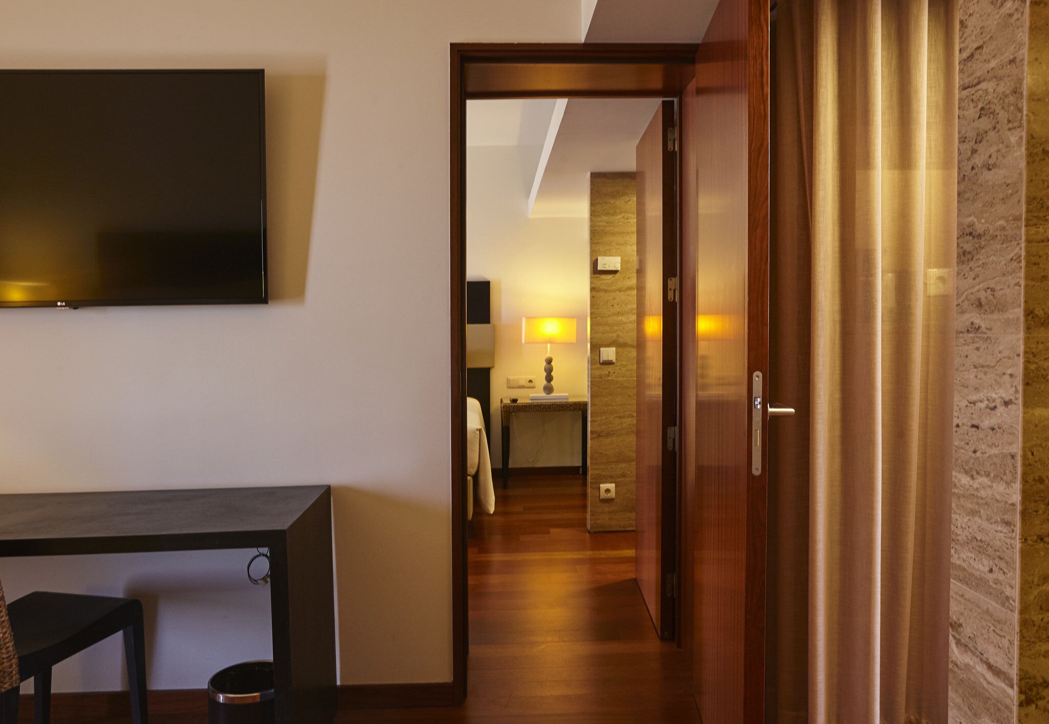 A hotel room with a flat-screen TV, a desk, and a chair. There is a wooden door leading to a bathroom with a sink and a mirror.