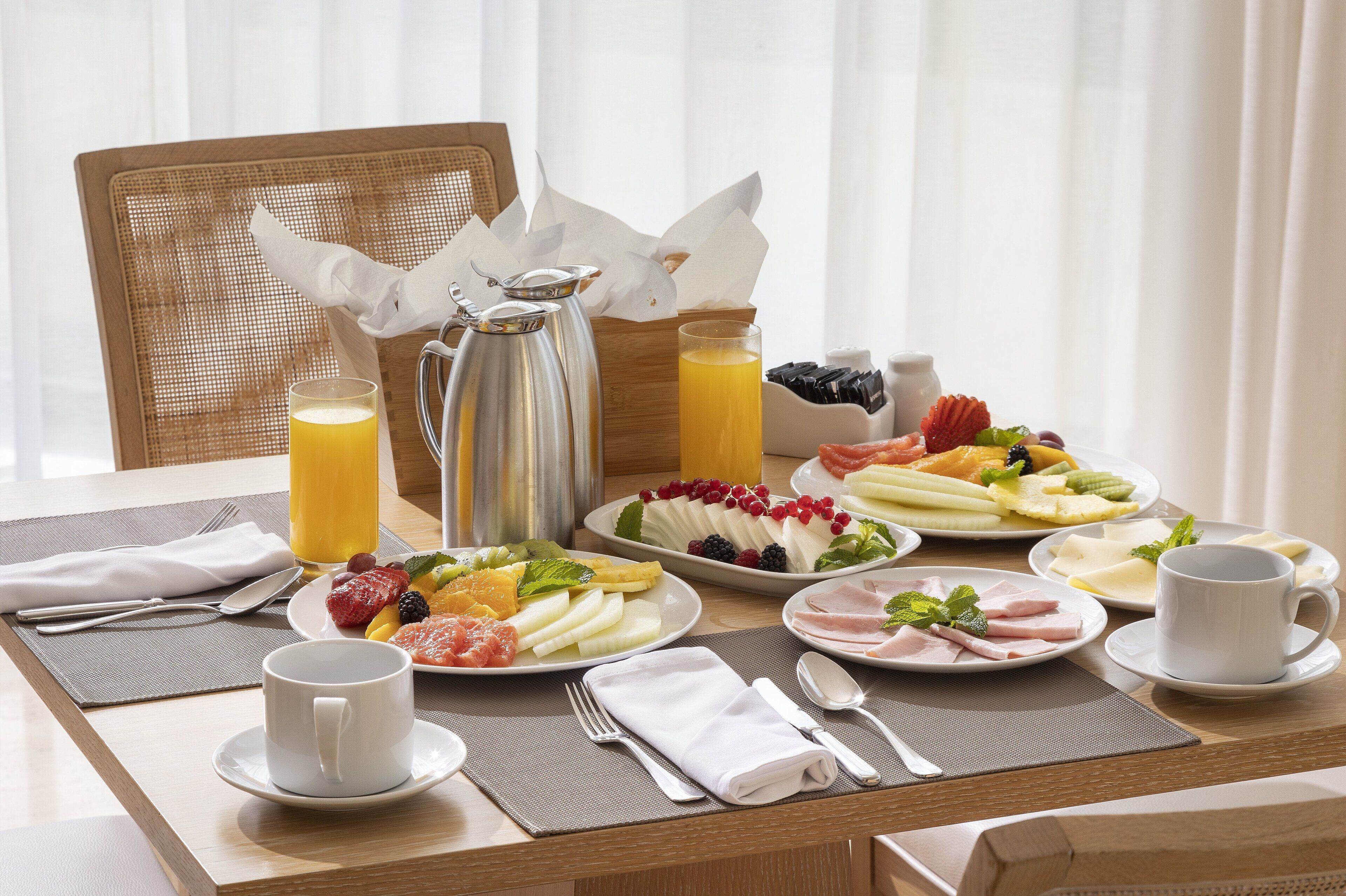 A breakfast table with plates of fruit, cheese, and ham, a coffee pot, and a glass of orange juice.