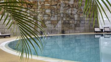 Una piscina interior con un mural de pared de piedra, sillas de descanso y una palmera.